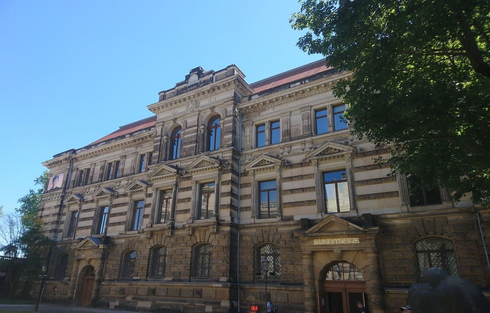 Прикрашений пісковиковий фасад Альбертінуму смугами світлого й темного каменю з вікнами під фронтонами та назвою над арковим входом, вигляд знизу, праворуч дерево.