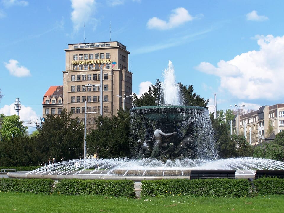 Kulatá kašna na Albertplatz s vodou stříkající přes bronzové postavy, vpředu živý plot a trávník a vzadu vysoká kancelářská věž pod modrou oblohou.