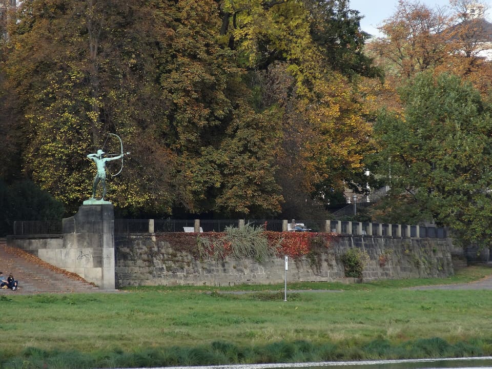 Malá socha lučištníka na vysokém kamenném pilíři na konci dlouhé pískovcové opěrné zdi, vlevo schody, vpředu trávník a vzadu stromy v podzimních barvách.