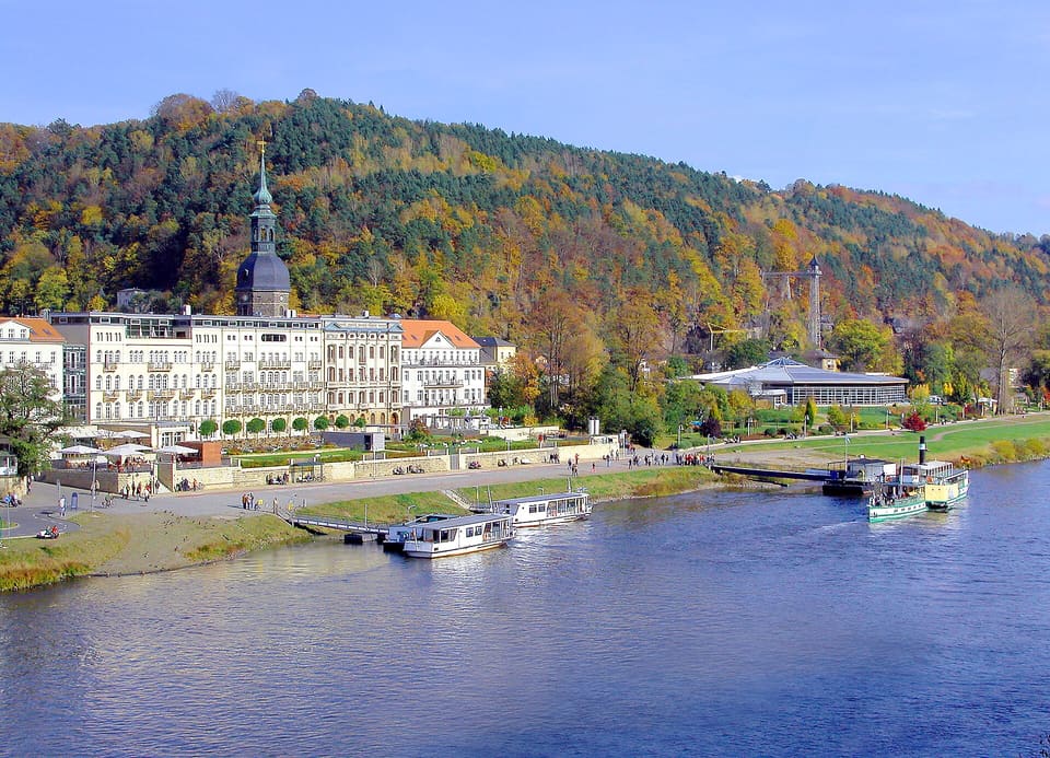 Nábřeží Bad Schandau s dlouhým průčelím hotelu a věží kostela pod podzimně zbarvenými zalesněnými kopci, u přístaviště na Labi lopatkové parníky.