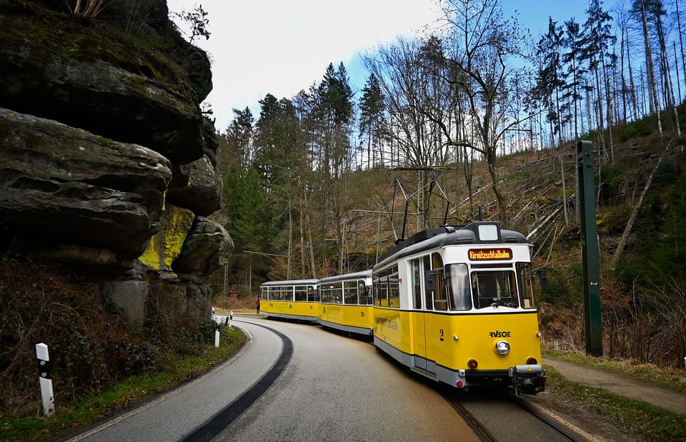 Žlutá tramvaj se dvěma přívěsnými vozy na zatáčející se silnici vedle skalního převisu, v lese holých stromů.
