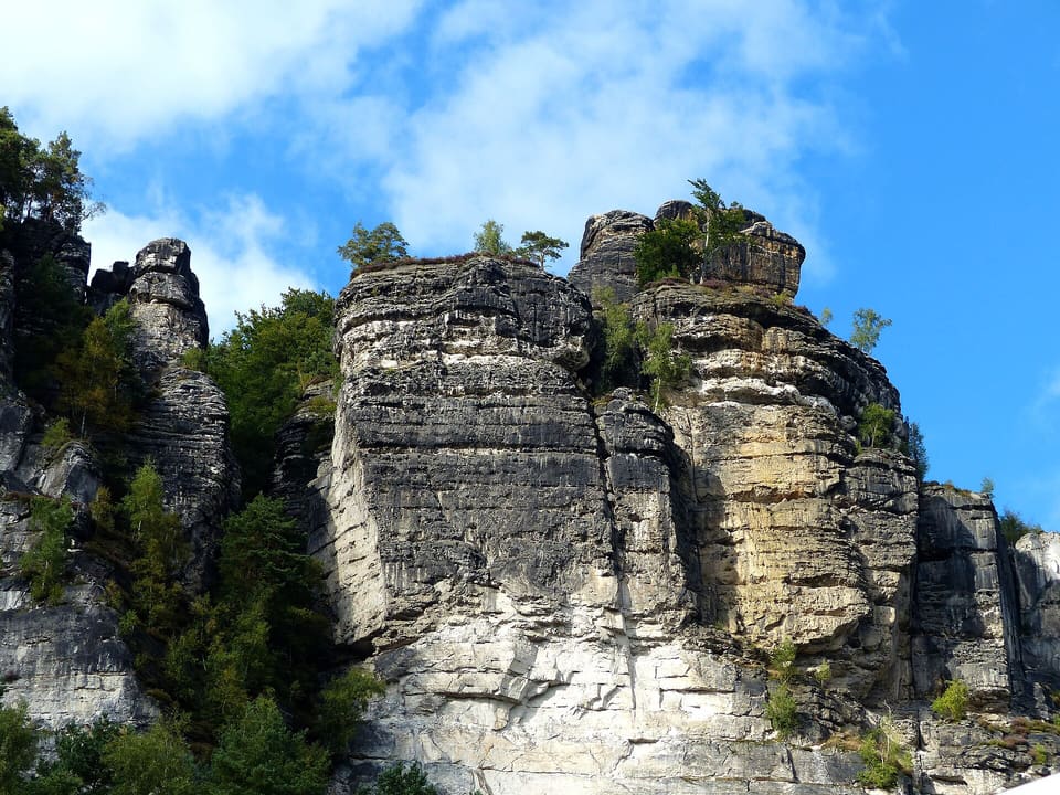 Pískovcové skalní stěny Bastei ve vodorovných vrstvách šedé a okrové barvy, zakončené borovicemi, pod modrou oblohou s tenkými bílými mraky.