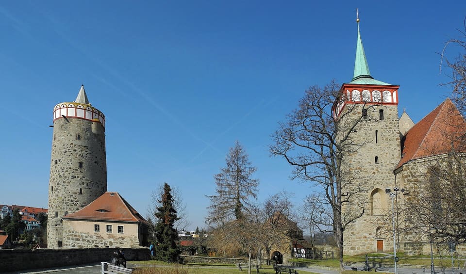 Kulatá kamenná věž s bílo-červenou korunou vlevo a čtyřhranná věž kostela Michaeliskirche se zelenou špicí vpravo pod sytě modrou oblohou.