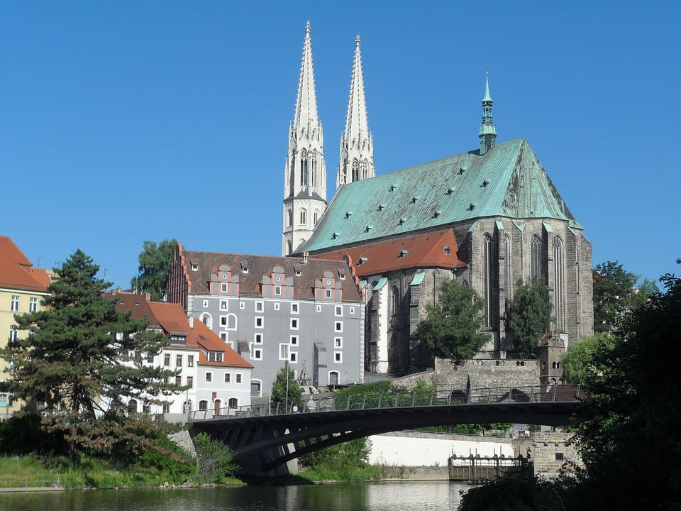 Dvojice věží a zelená měděná střecha kostela Peterskirche nad mostem přes řeku Nisu, pod jasně modrou oblohou.