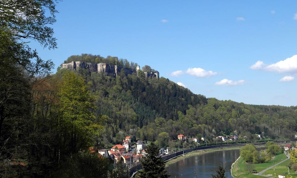 Pevnost Königstein na okraji zalesněné stolové hory nad Labem, dole řeka a vesnice Königstein, pod modrou oblohou.