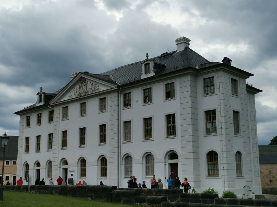 Brunnenhaus, dlouhá bílá budova s tympanonem uprostřed a řadami obloukových oken, pod tmavou zataženou oblohou, před ní několik návštěvníků.