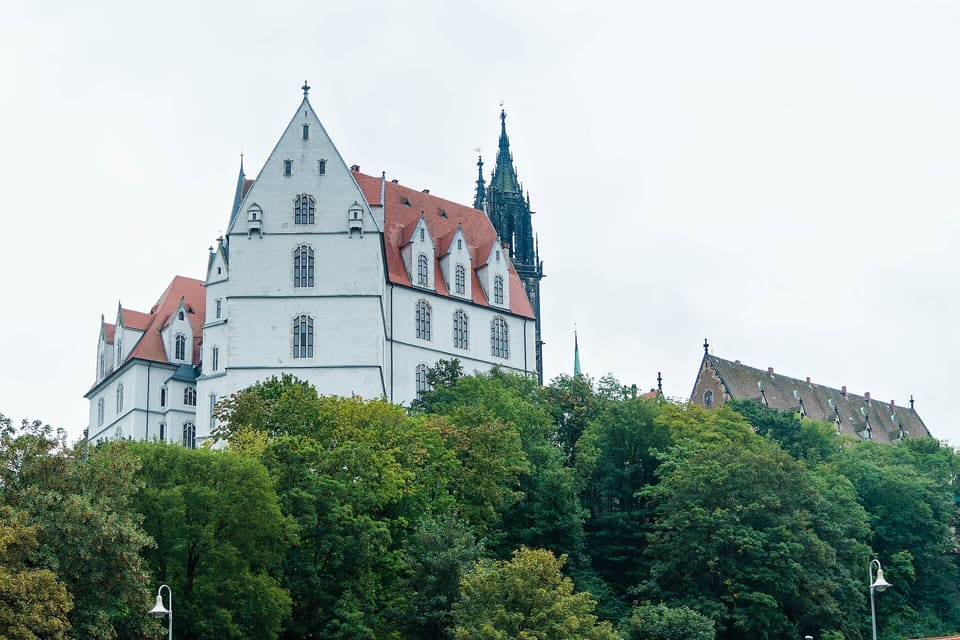 Bílá štítová zeď Albrechtsburgu a červená střecha nad zelenými stromy, vpravo se proti bledé obloze zvedá prolamovaná věž katedrály.
