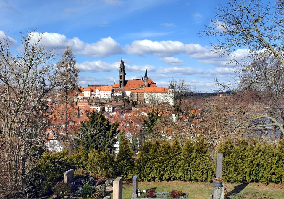Hradní vrch v Míšni nad domy s červenými střechami a hřbitovem, na vrcholu věž katedrály a Albrechtsburg, pod modrou oblohou s bílými mraky a holými stromy rámujícími pohled.