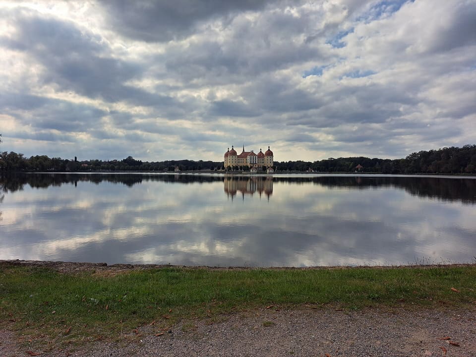 Schloss Moritzburg na protějším břehu klidného jezera, malý zámek s věžemi odrážejícími se ve vodě, pod těžkou oblohou šedých a bílých mraků.