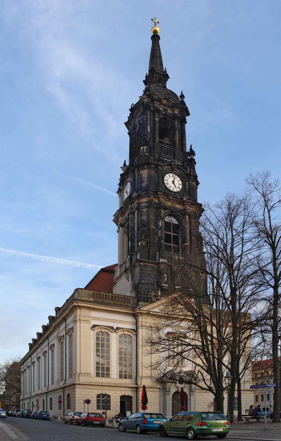 Ciemna piaskowcowa wieża kościoła Dreikönigskirche z tarczą zegara i pozłacanym szczytem wznosząca się nad jasną bryłą kościoła, z nagimi drzewami i zaparkowanymi samochodami wzdłuż ulicy z przodu.
