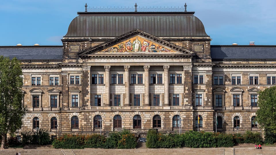 The central part of a stone building with a portico of six columns and a pediment filled with a colourful picture, below a dome-shaped roof.