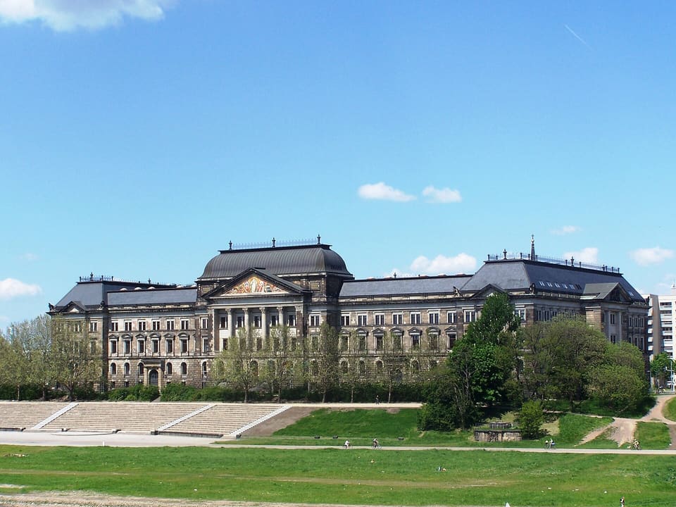 A long stone building seen at an angle from the river meadow, with a pedimented centre under a dome-shaped roof, wide steps at the left and trees in front.