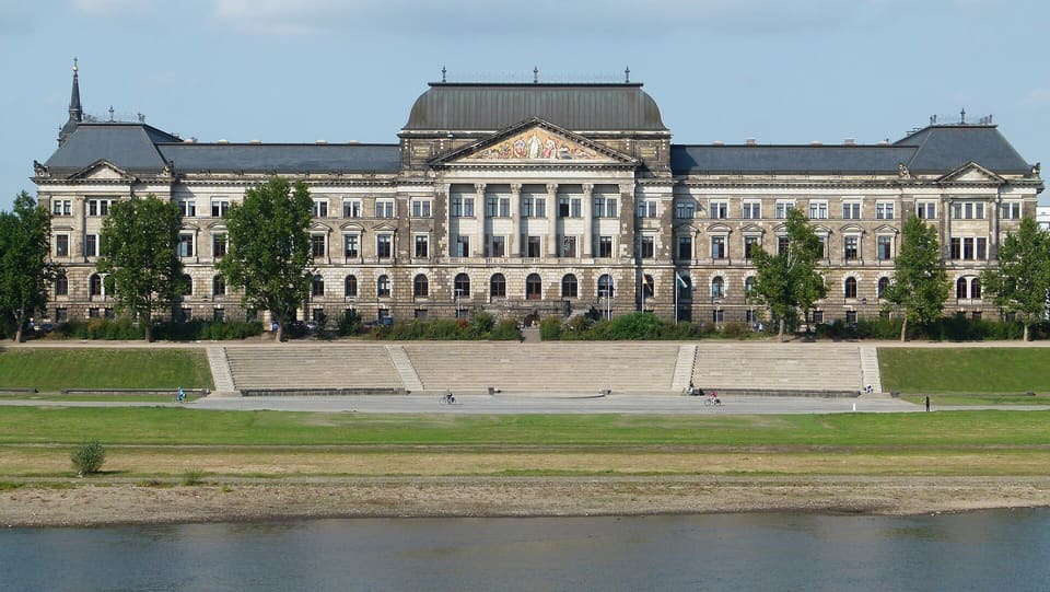 A long neoclassical building in pale and dark stone with a central portico and pediment under a dome-shaped roof, above a wide flight of stone steps and a grassy river bank.