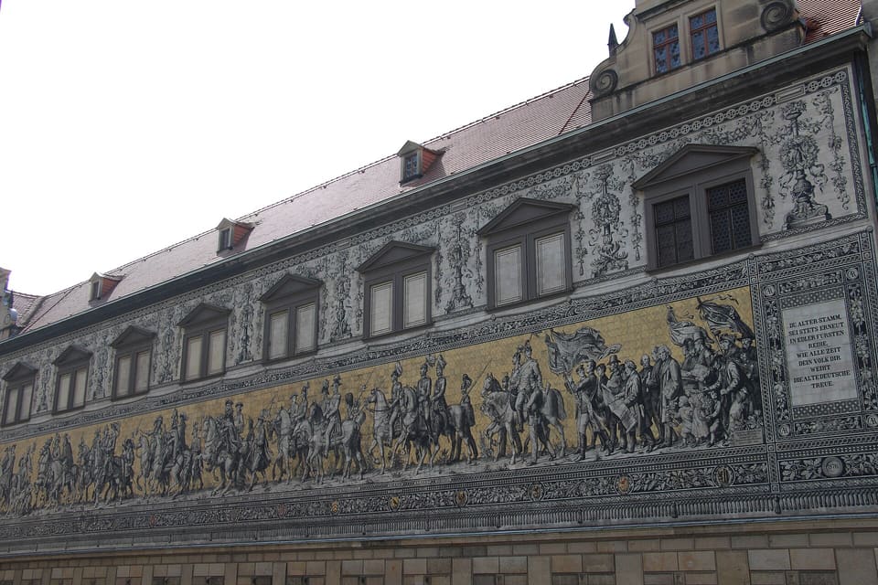 The Fürstenzug, a long frieze of mounted rulers in dark figures on gold tiles, with an inscription panel at the right, seen at an angle along its wall.
