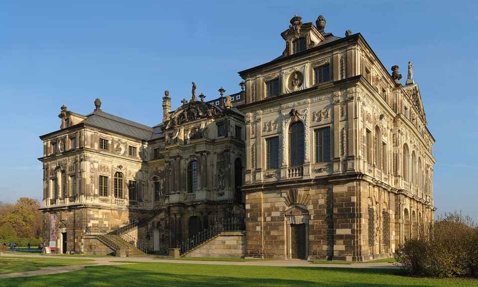 Bogaty barokowy piaskowcowy front Palais im Großen Garten z rzeźbionymi ozdobami, stromymi dachami i wysokimi oknami, widziany przez trawnik pod bezchmurnym błękitnym niebem.