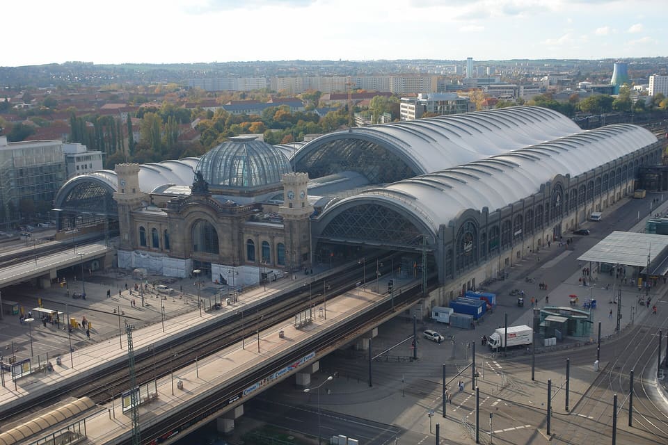 Długa, srebrzysta hala peronowa Hauptbahnhof z dachem membranowym i piaskowcowy budynek wejściowy z bliźniaczymi wieżami zegarowymi i szklaną kopułą, widziane z góry przez tory i plac przed dworcem.