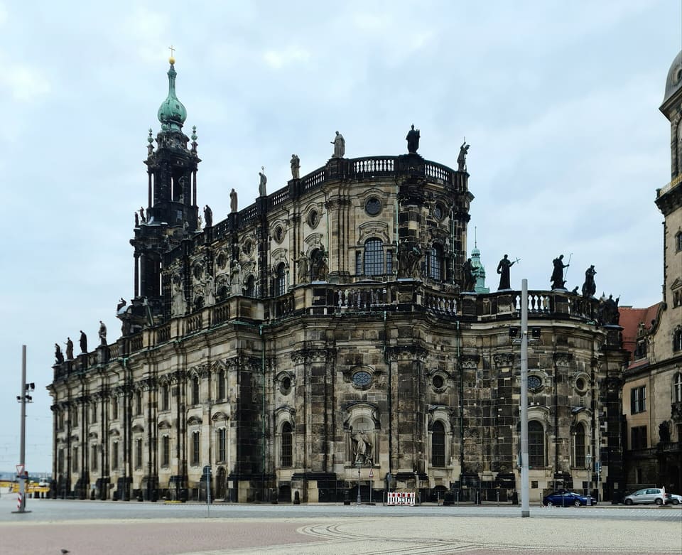 The Hofkirche's dark Baroque nave and apse with statues along the balustrade, and its green-capped tower at the left, seen across the square on a cloudy day.