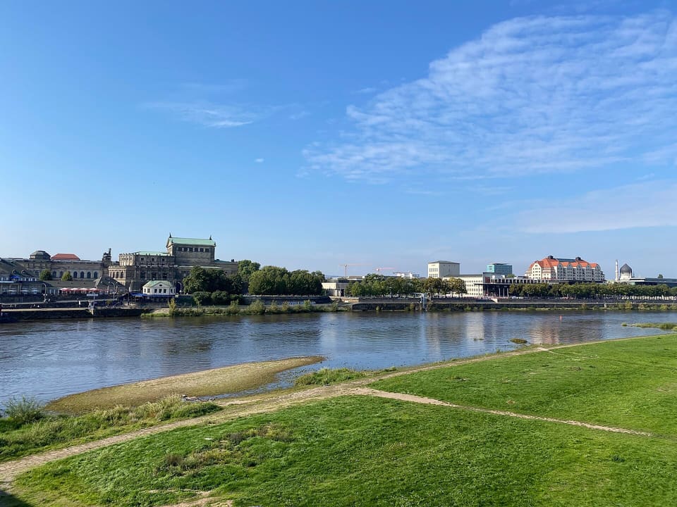 Ein grasbewachsenes Flussufer und eine Sandzunge an der Elbe, am anderen Ufer eine niedrige Gebäudesilhouette unter blauem Himmel mit dünnen Wolken.