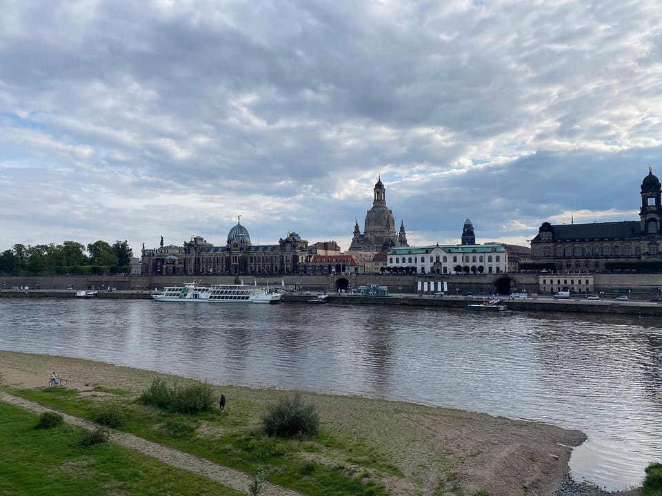 Die Elbe unter bewölktem Himmel mit einer sandigen Uferbank im Vordergrund und am anderen Ufer einer langen Gebäudereihe, einer Kuppelkirche, einem Turm und einem weißen Flussschiff am Kai.