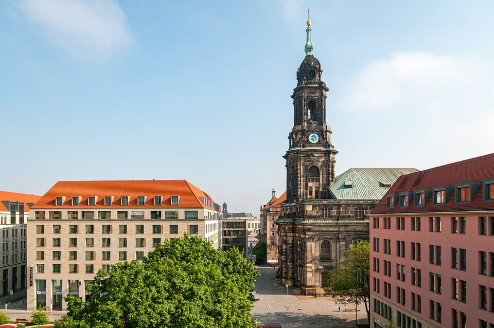 Ciemna piaskowcowa wieża Kreuzkirche z zielonym miedzianym hełmem i tarczami zegarowymi ponad dachem kościoła, między czerwonym a różowym budynkiem, widziana z góry.
