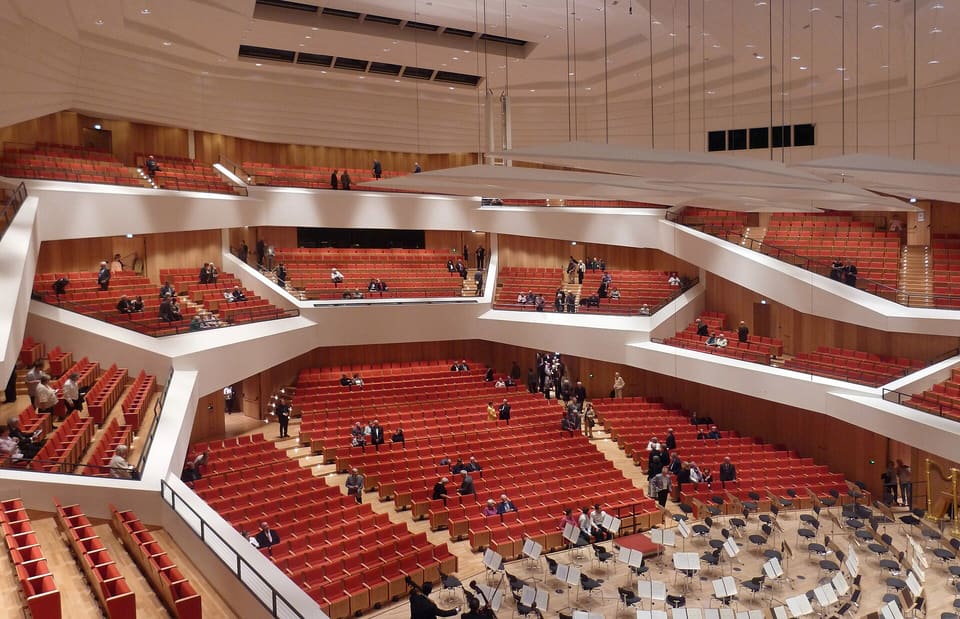 Sala koncertowa widziana z balkonu, z tarasowo ułożonymi blokami czerwonych foteli, białymi zakrzywionymi frontami balkonów, ciepłymi drewnianymi okładzinami ścian, zawieszonymi białymi reflektorami i pulpitami na scenie poniżej.