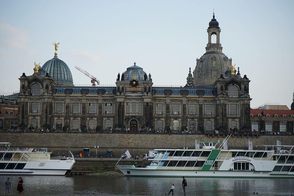 The Kunstakademie's long facade with its glass dome and the Frauenkirche's dome beyond, seen across the Elbe above the embankment wall with two river steamers moored in front.