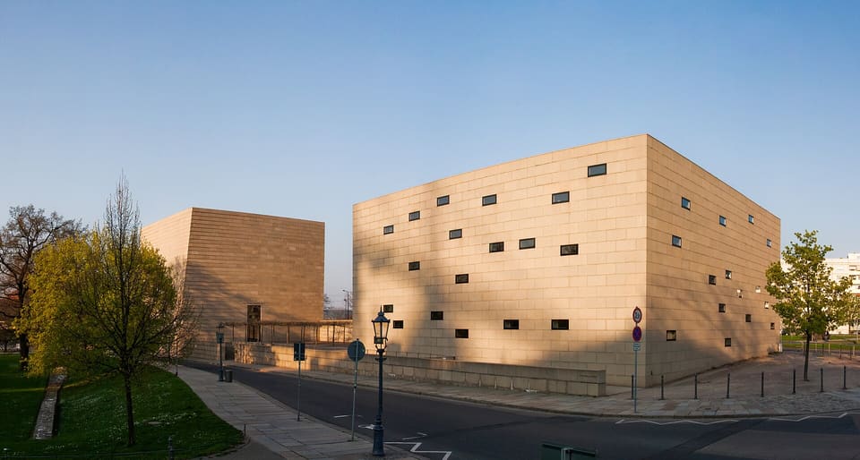Dvě prosté krychle ze světlého, světle hnědého kamene Neue Synagoge, větší s roztroušenými malými okenními štěrbinami, na rohu ulice s mladými stromy a plynovou lampou pod jasnou oblohou.