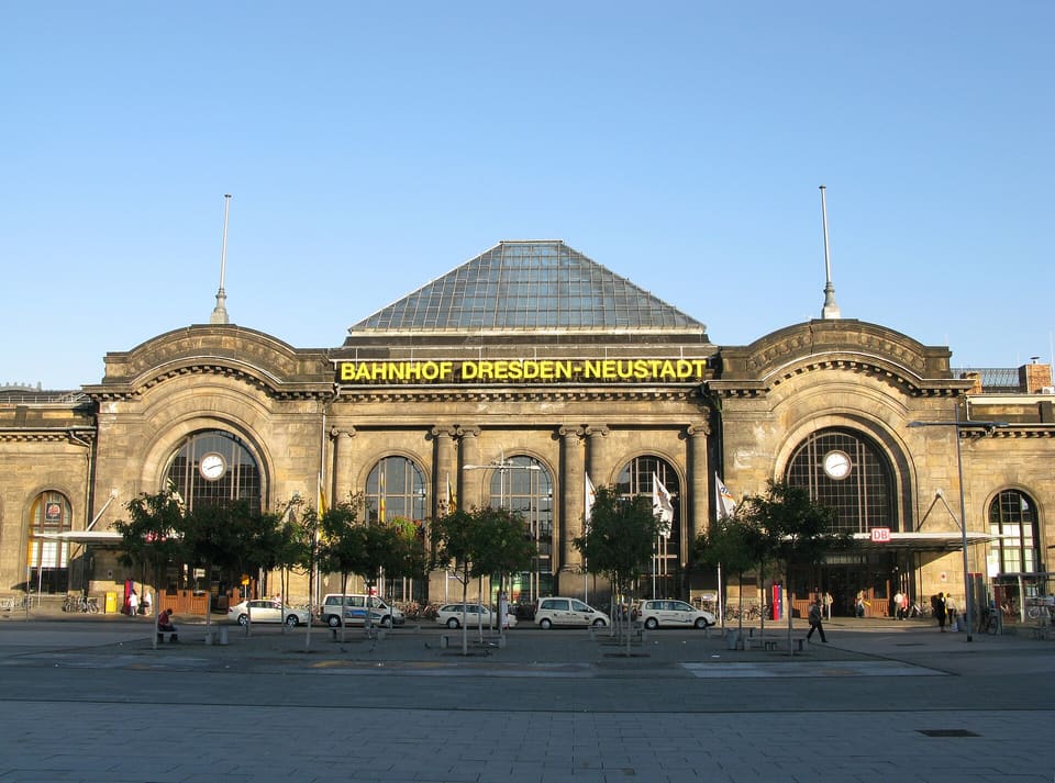 Pískovcová výpravní budova nádraží Neustadt s velkými obloukovými okny, kulatými hodinami, střechou ve tvaru skleněné pyramidy a nápisem Bahnhof Dresden-Neustadt, při pohledu z předprostoru pod jasně modrou oblohou.
