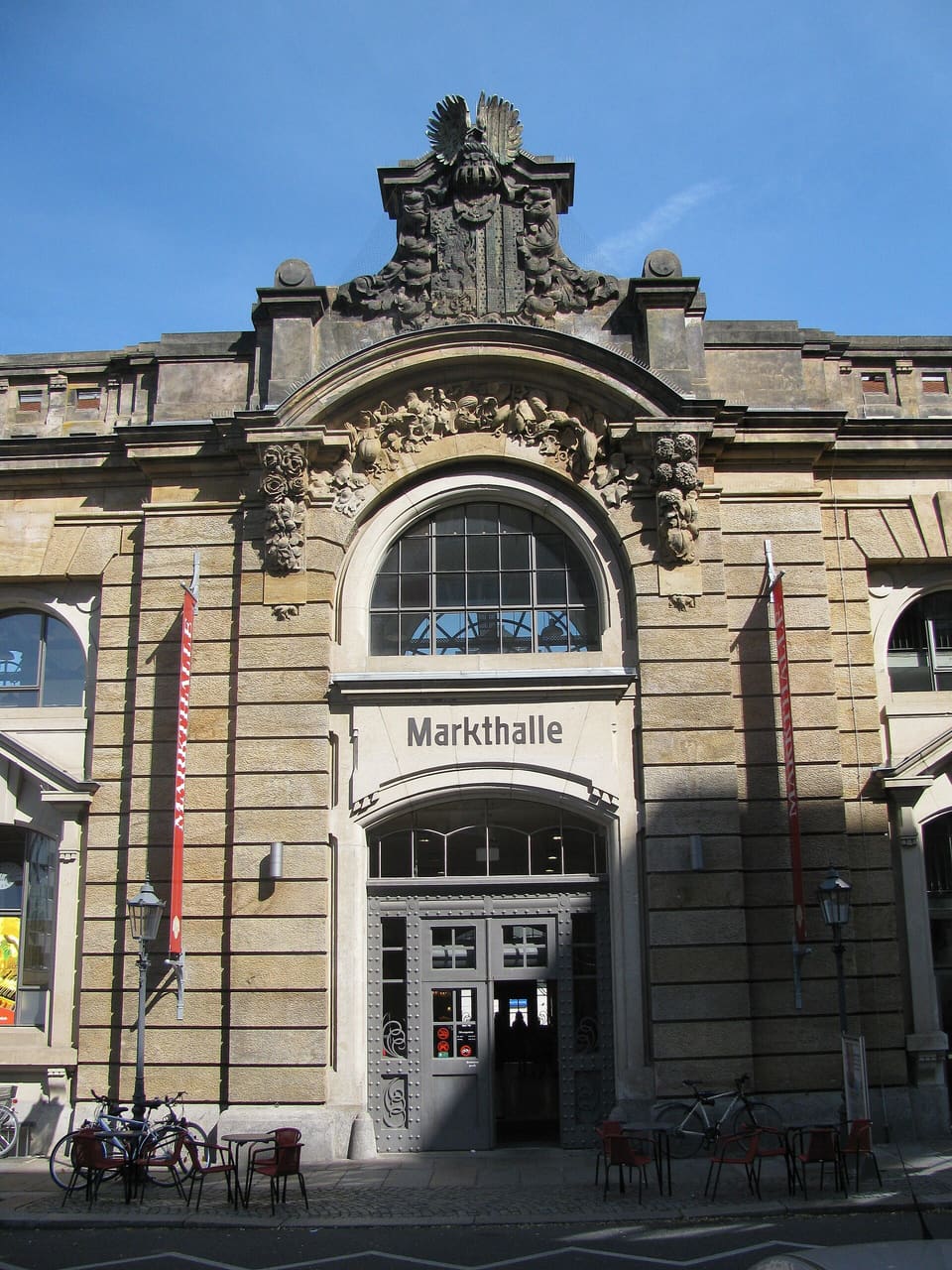 Pískovcové vstupní průčelí Neustädter Markthalle s velkým obloukovým oknem, vyřezávaným znakem nahoře a nápisem Markthalle nad dveřmi, na úrovni ulice kavárenské židle a kola.
