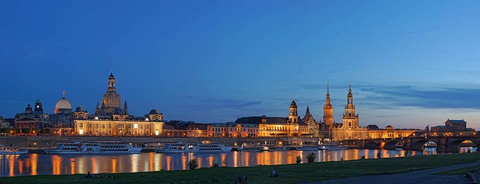 The Altstadt across the Elbe at dusk under a deep blue sky: the glass dome of the Kunstakademie and the dome of the Frauenkirche at the left, the lit Hofkirche tower and the Hausmannsturm of the Residenzschloss at the centre, the Augustus Bridge at the right, and river boats along the quay, all reflected in the water.