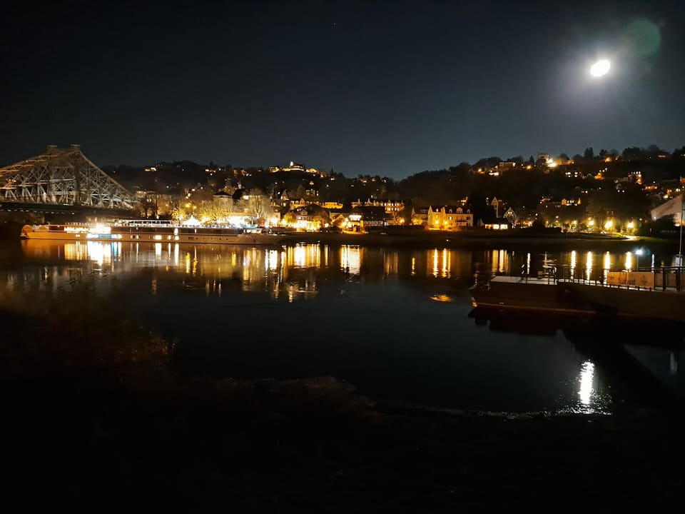 The steel truss of the Blaues Wunder at the left edge, a moored boat and lit houses on the wooded hillside above the Elbe, a bright moon in the sky and the lights reflected in the dark river.