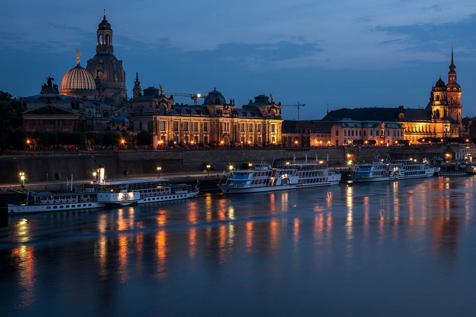 The Kunstakademie's glass dome with the Frauenkirche's dome behind it at the left and the lit Hausmannsturm at the right, above the Terrassenufer's wall and river boats, in the blue hour after sunset.