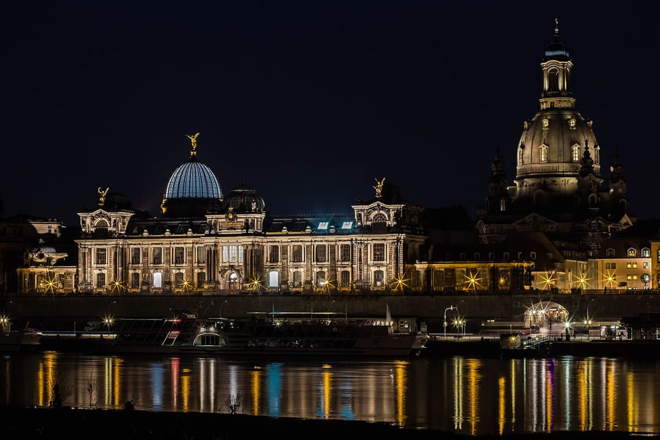 The Kunstakademie with its glass dome and the lit dome of the Frauenkirche above the Elbe's far bank, with the lights reflected in the river in the foreground.