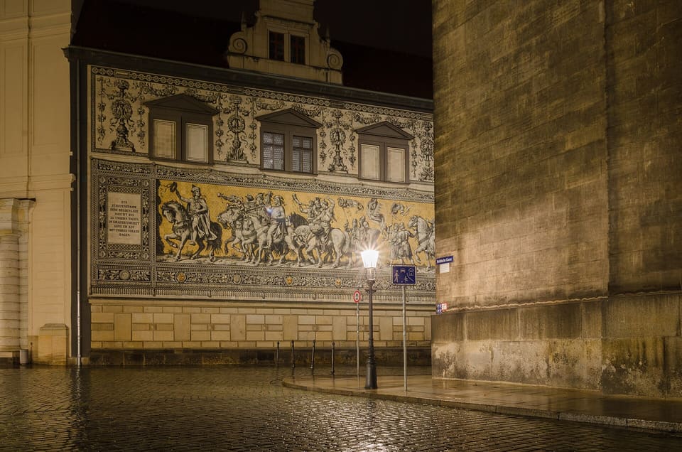Part of the Fürstenzug's tile mural of mounted riders, lit at night above a wet cobbled street with one street lamp in front and the sandstone wall of the Hofkirche at the right.