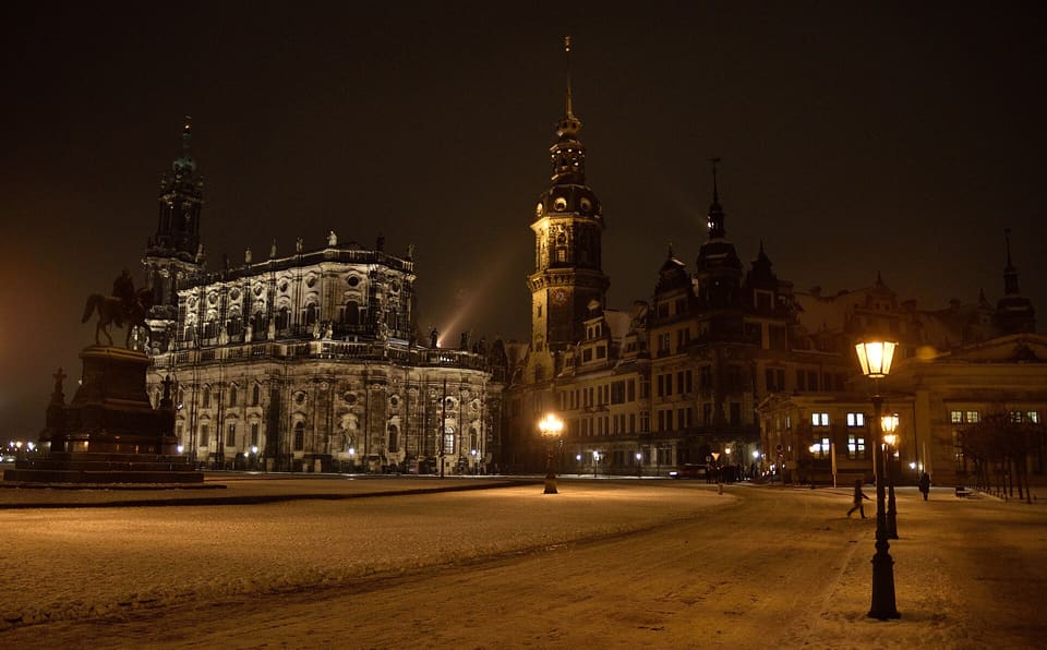 The Hofkirche at the left and the Residenzschloss with its tall Hausmannsturm at the right, seen across a snow-covered Theaterplatz at night, with the equestrian statue at the left edge and a lit street lamp in front.