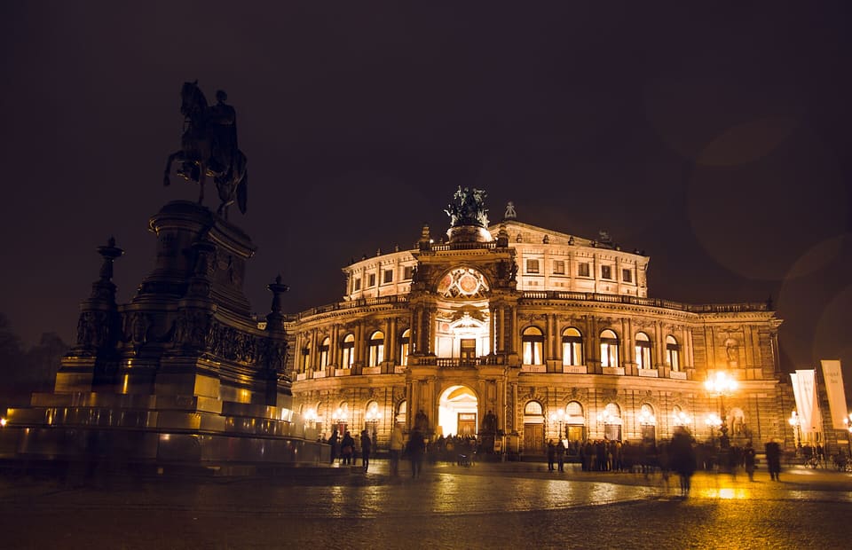 The Semperoper's lit front in the evening, with the equestrian statue of King John on its pedestal at the left, people at the entrance and a wet square reflecting the lamps.