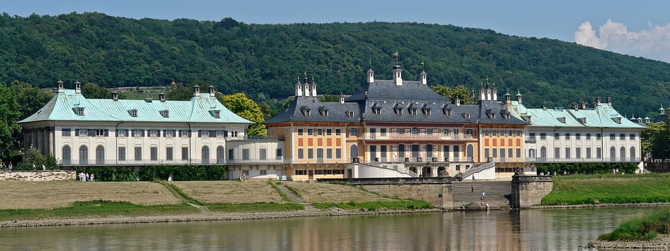 Палац Пільніц з боку Ельби: довгі світлі крила із зеленими мідними дахами обабіч центрального блоку з темним шатровим дахом, сходи до річки та лісисті пагорби позаду.