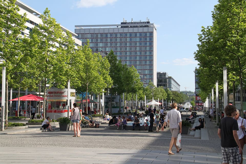 Pěší Prager Straße za slunečného dne, lemovaná mladými stromy, lidé chodí a sedí pod slunečníky a vzadu se zvedá vysoká hotelová věž.