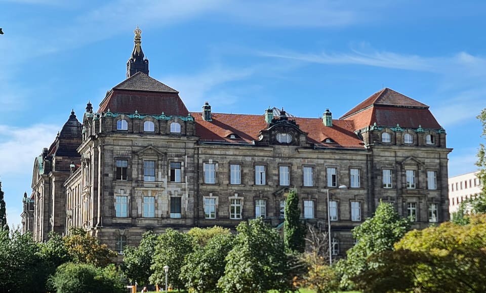 The dark sandstone side of a large building with red roofs, a domed tower with a gilded finial and rows of tall windows, framed by green trees.