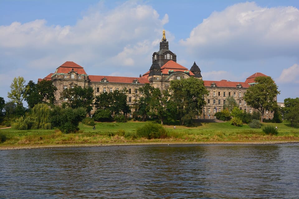 A wide sandstone building with red roofs and a central domed tower behind willows and a grassy bank, seen from the water under a blue sky with clouds.