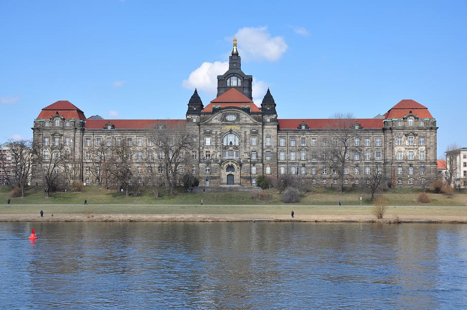 A long sandstone building with red-tiled wings and a central tower under a domed roof, seen across the Elbe and a strip of lawn under a blue sky with white clouds.