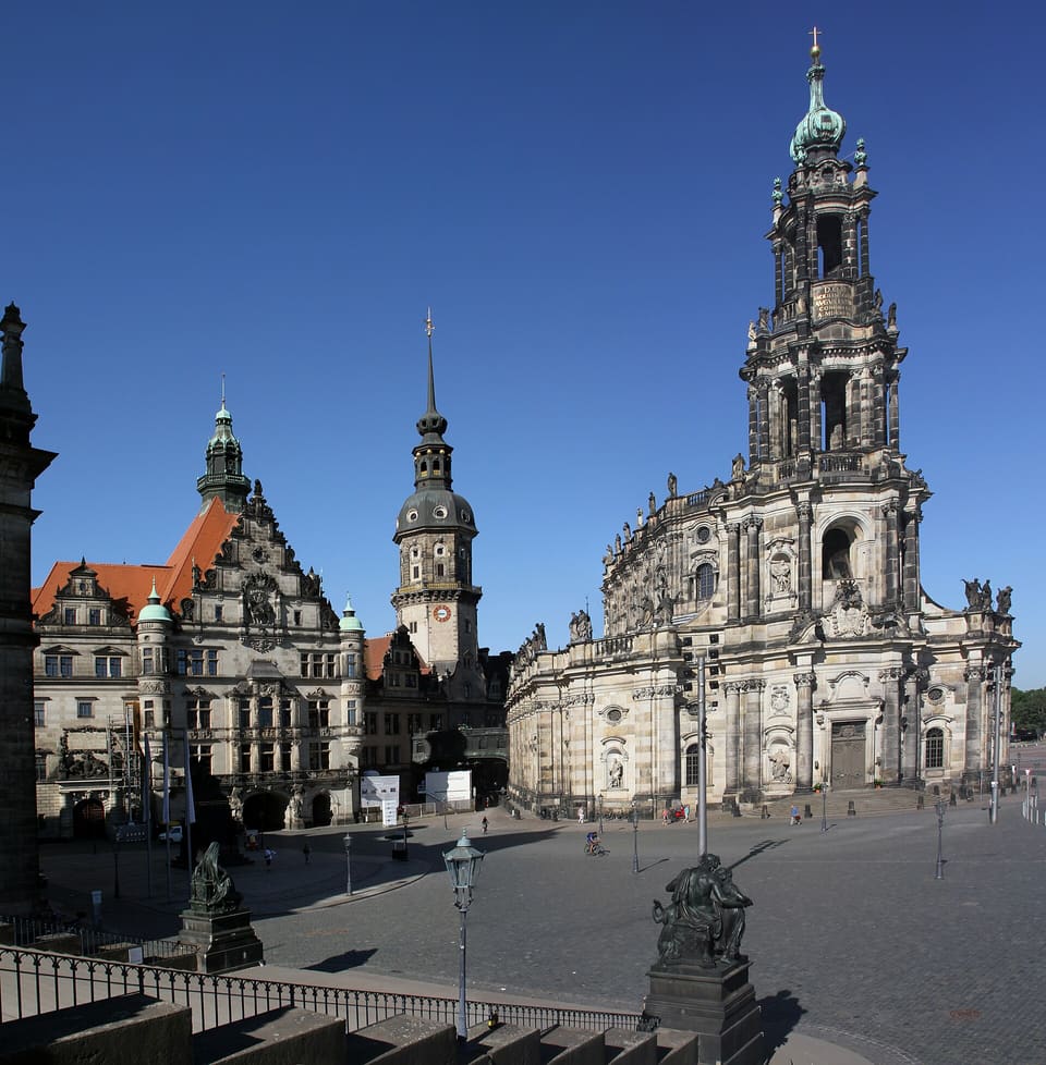 Vysoká barokní věž kostela Hofkirche se zelenou čepicí a světlá pískovcová bočnice vedle zdobeného křídla Georgenbau s červenou střechou a věže Hausmannsturm, přes dlážděný Schlossplatz pod sytě modrou oblohou.
