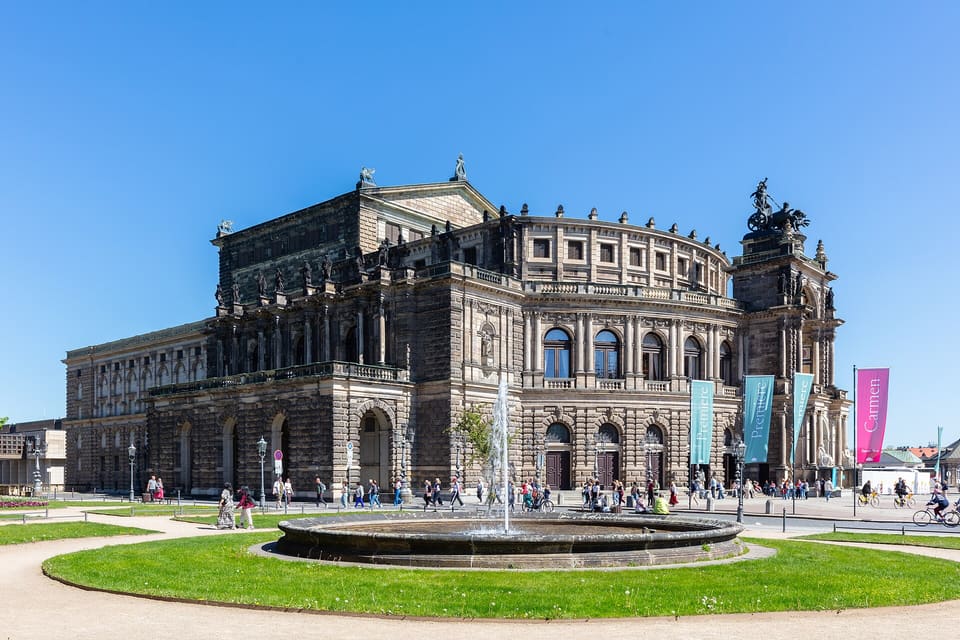The Semperoper's curved sandstone front seen across a lawn and a fountain basin, under a clear blue sky.