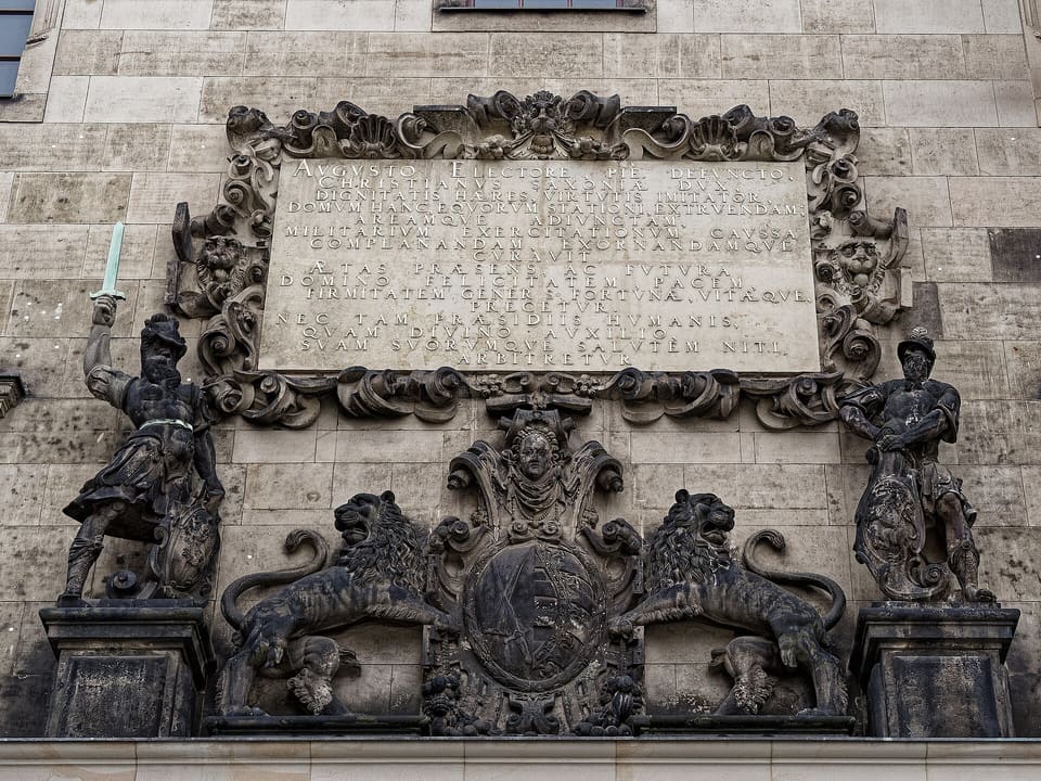 Tablica z napisem w ozdobnej ramie zwojowej nad herbem zwieńczonym małym koronowanym popiersiem, z ciemnym kamiennym lwem po każdej stronie i na zewnętrznych końcach dwiema ciemnymi postaciami w zbrojach, lewą unoszącą miecz, wszystko na jasnej piaskowcowej ścianie.