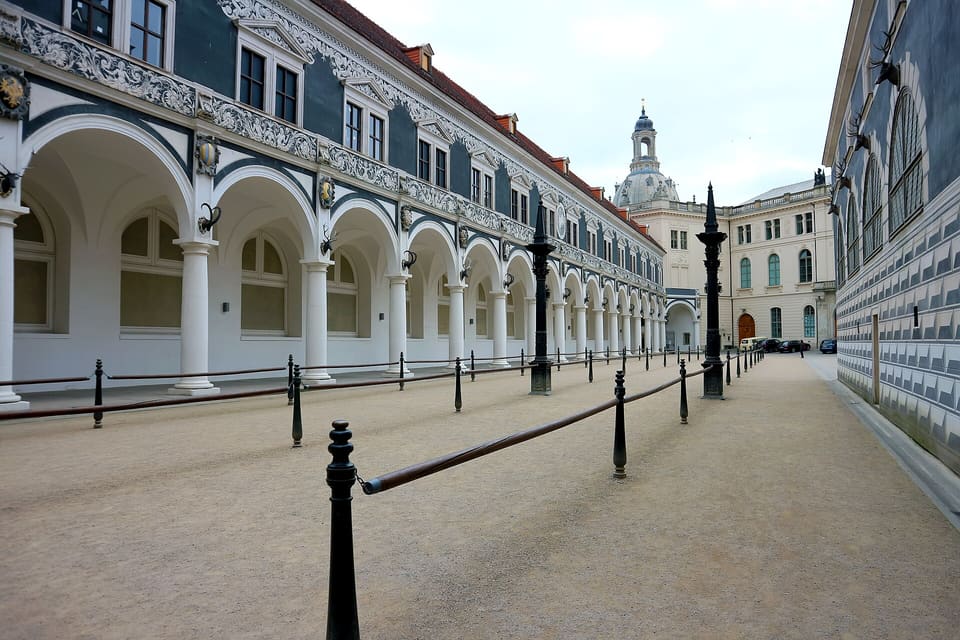 Długie podcienie z białych kolumn i okrągłych łuków pod bogato zdobioną ciemnoniebieską górną ścianą Stallhof, z rzędem czarnych słupków wzdłuż żwirowego dziedzińca i budynkiem z kopułą w oddali.