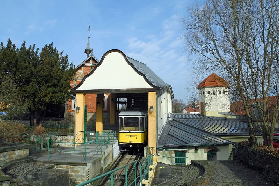 Žlutý vůz pozemní lanovky vyjíždějící z oblé štítové stanice na vrcholu trati, kolem červená cihlová věž a holé stromy pod modrou oblohou.