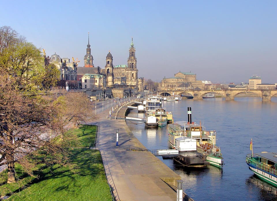 Dlážděná promenáda u Labe v teplém světle se zakotvenými lopatkovými parníky, vlevo holý strom a v dálce věže a dlouhý most.
