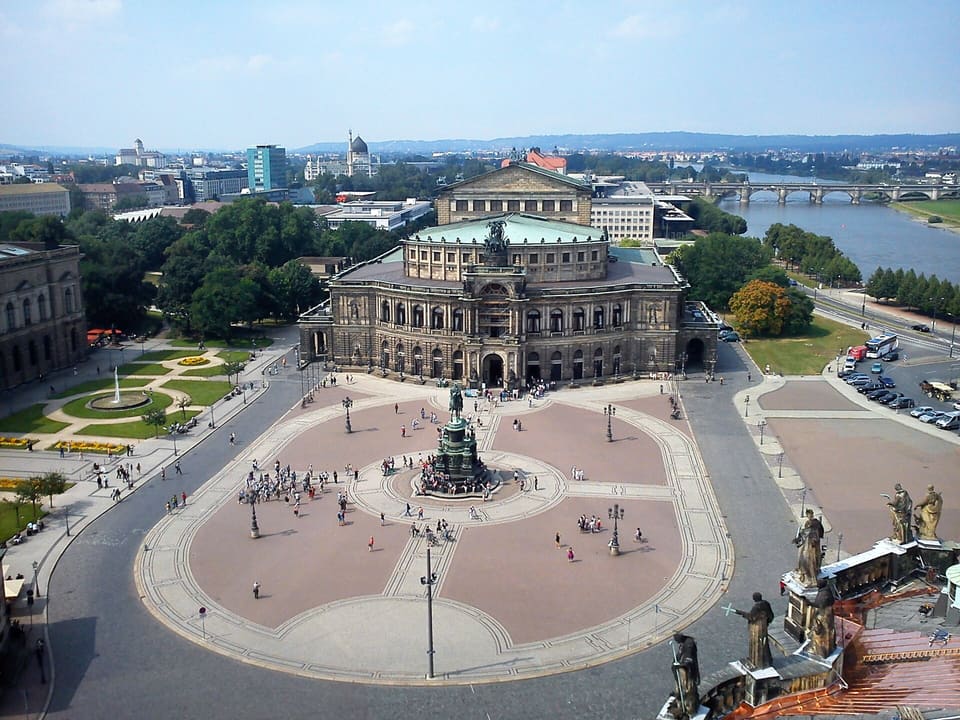 Zelenou střechou krytý pískovcový blok Semperoper za oválným, růžově dlážděným Theaterplatz s jezdeckým pomníkem uprostřed, vlevo trávníky a stromy a za nimi Labe s mostem.