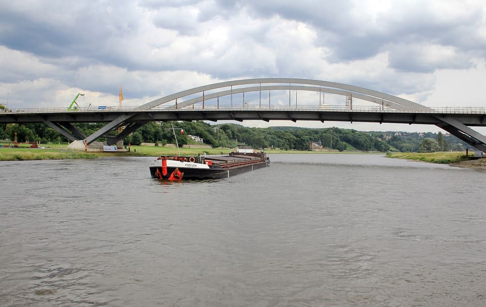 Štíhlý bílý ocelový oblouk mostu Waldschlösschenbrücke přes široké šedé Labe, pod ním proplouvá nákladní člun, za ním zelené břehy.