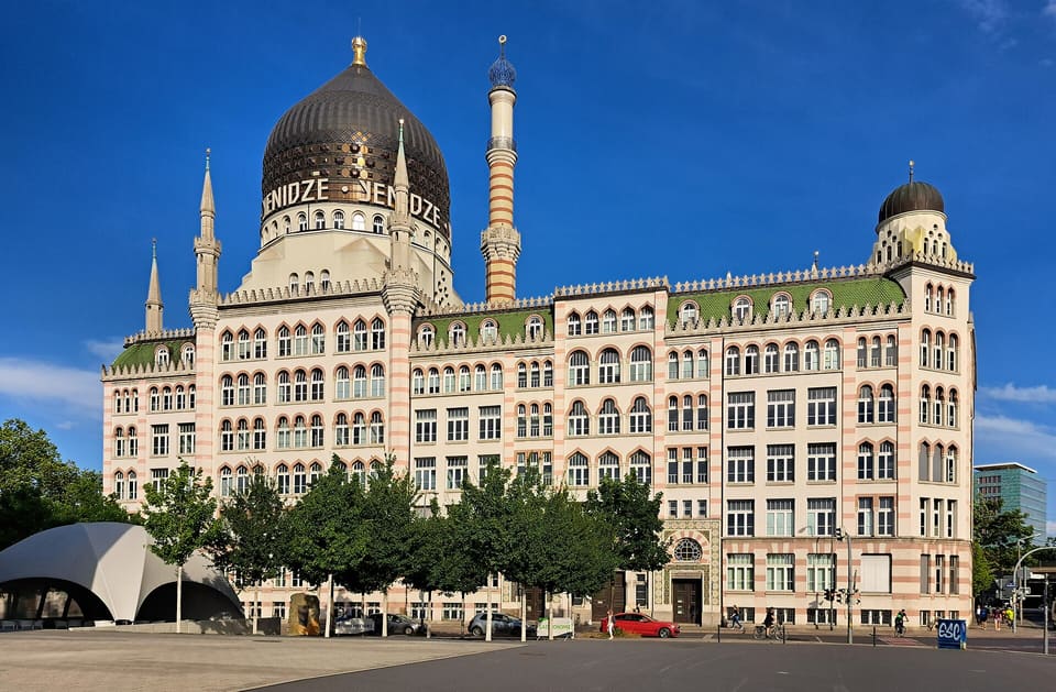 The Yenidze, a former cigarette factory built to look like a mosque, with its dark dome, striped minaret and green-roofed ranges behind a row of young trees under a deep blue sky.